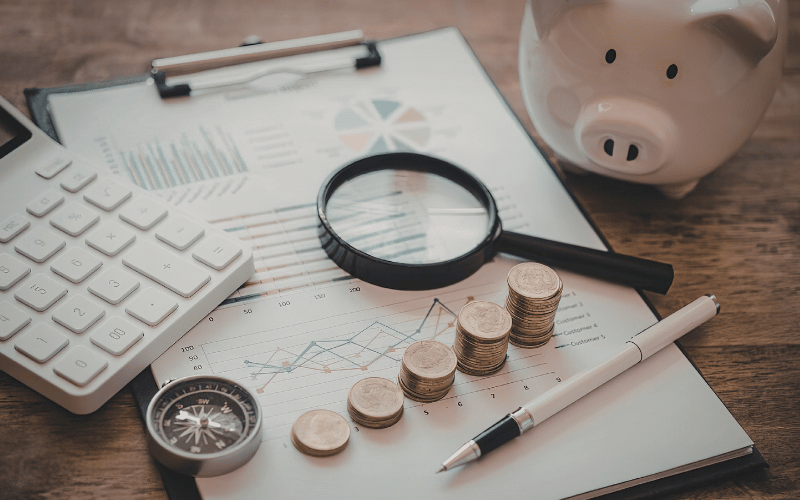 financial paperwork on a desk with coins, calculator, magnify glass, compass, pen, and a piggy bank